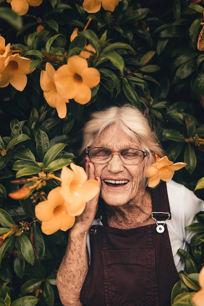 San Diego Notary, an older woman is partially standing in a large bush, she smiles as the flowers surround her